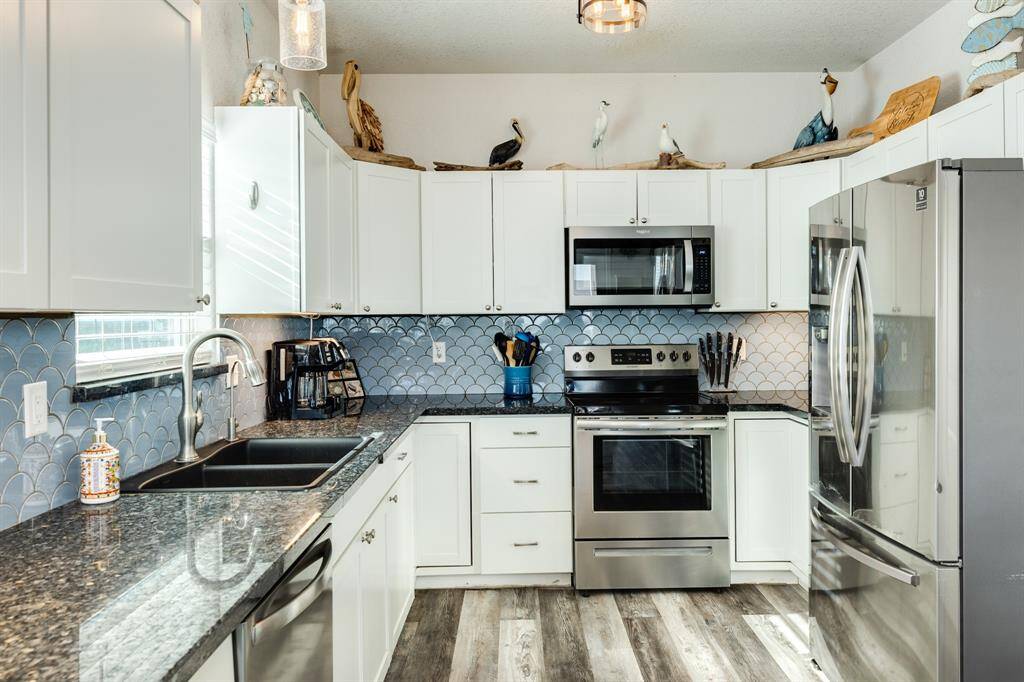 This modern kitchen features white cabinetry, stainless steel appliances, and a stylish blue tile backsplash. The granite countertops and wood-look flooring add a touch of elegance, while ample natural light brightens the space.