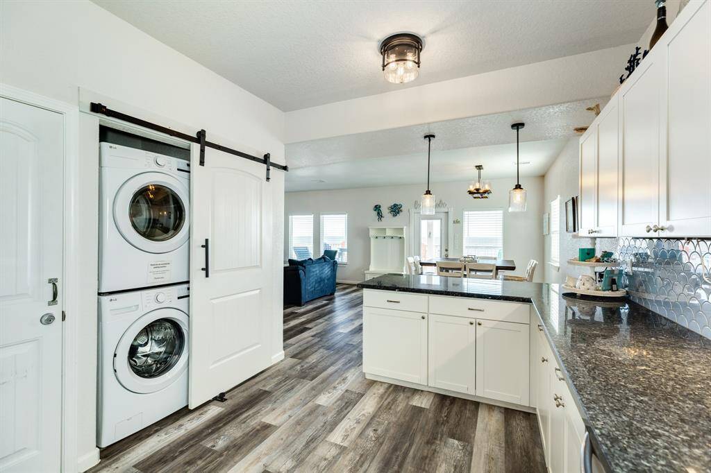 Bright, open kitchen with modern white cabinetry and granite countertops. Features a stacked washer-dryer neatly tucked behind a sliding barn door. The space flows into a dining and living area with natural light.