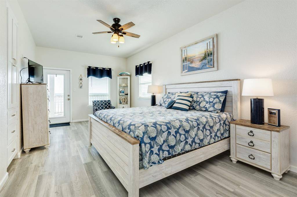 Bright and airy 2nd floor primary bedroom with light flooring, a cozy bed featuring blue floral bedding, and matching nightstands. The room includes a ceiling fan, a wall-mounted TV, and deck access through a glass door, creating a welcoming and comfortable space.