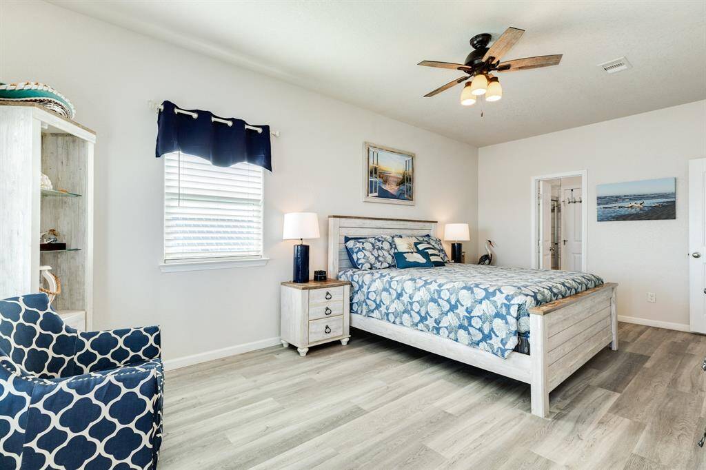 This photo showcases a bright and airy 2nd floor primary bedroom with modern decor. It features a comfortable bed with patterned bedding, a ceiling fan, and wood-style flooring. A cozy armchair and stylish nightstands add to the inviting atmosphere, while large windows provide plenty of natural light.