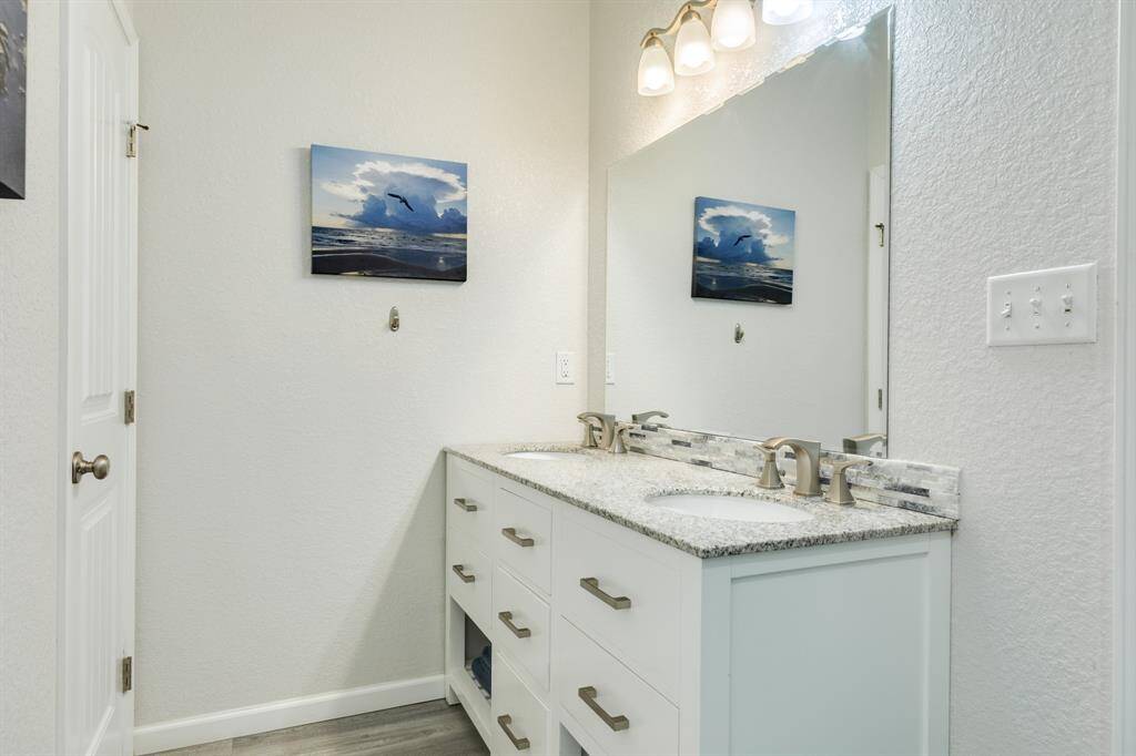 This 2nd floor primary bathroom features a modern double vanity with granite countertops, sleek faucets, and ample drawer storage. The space is brightened by overhead lighting and a large mirror, complemented by a serene ocean-themed wall art.