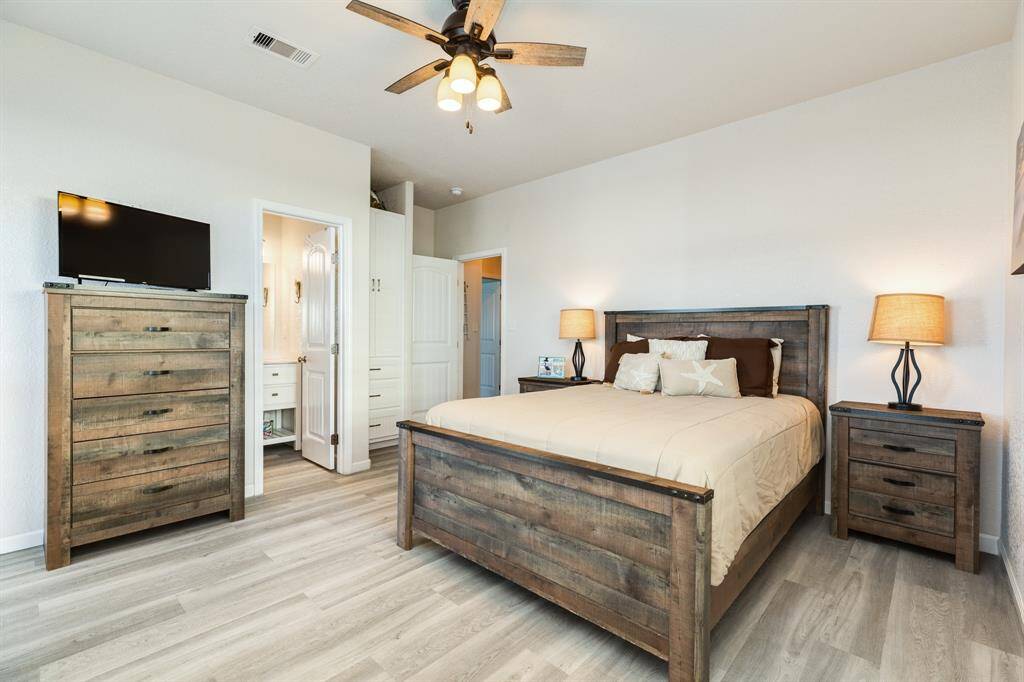 The 3rd bedroom features a rustic wooden bed frame, matching nightstands, and a tall dresser. It includes a ceiling fan, neutral tones, and an attached bathroom. The room is well-lit with two lamps, and there's a mounted TV for entertainment.