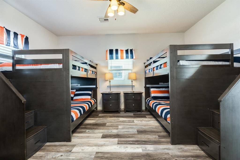 This 1st floor bedroom features two sets of dark wood bunk beds with built-in stairs. The room is brightened by a ceiling fan light and two windows with colorful striped curtains. A central nightstand with lamps offers additional lighting, and the wood-look flooring adds warmth. Perfect for a family or guest room as it comes with an ensuite.