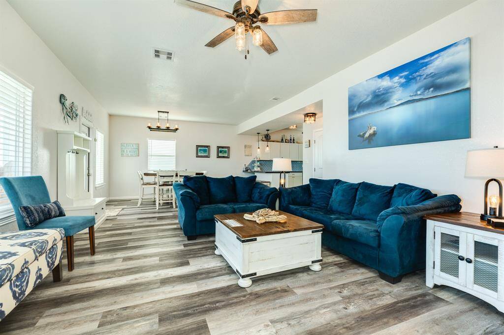 This inviting living room features modern decor with a cozy blue sectional sofa, stylish wood flooring, and ample natural light. An open layout connects to the dining area and kitchen, enhanced by a ceiling fan and tasteful wall art.