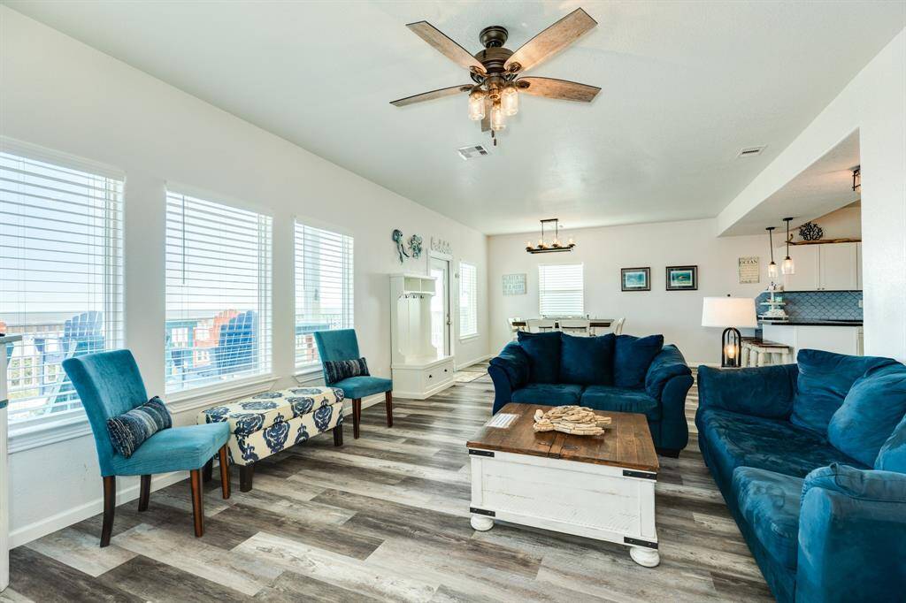Bright and inviting living space with large windows, modern ceiling fan, and stylish flooring. The room features plush blue sofas, a cozy dining area, and viewsof the open-concept kitchen and dining area.
