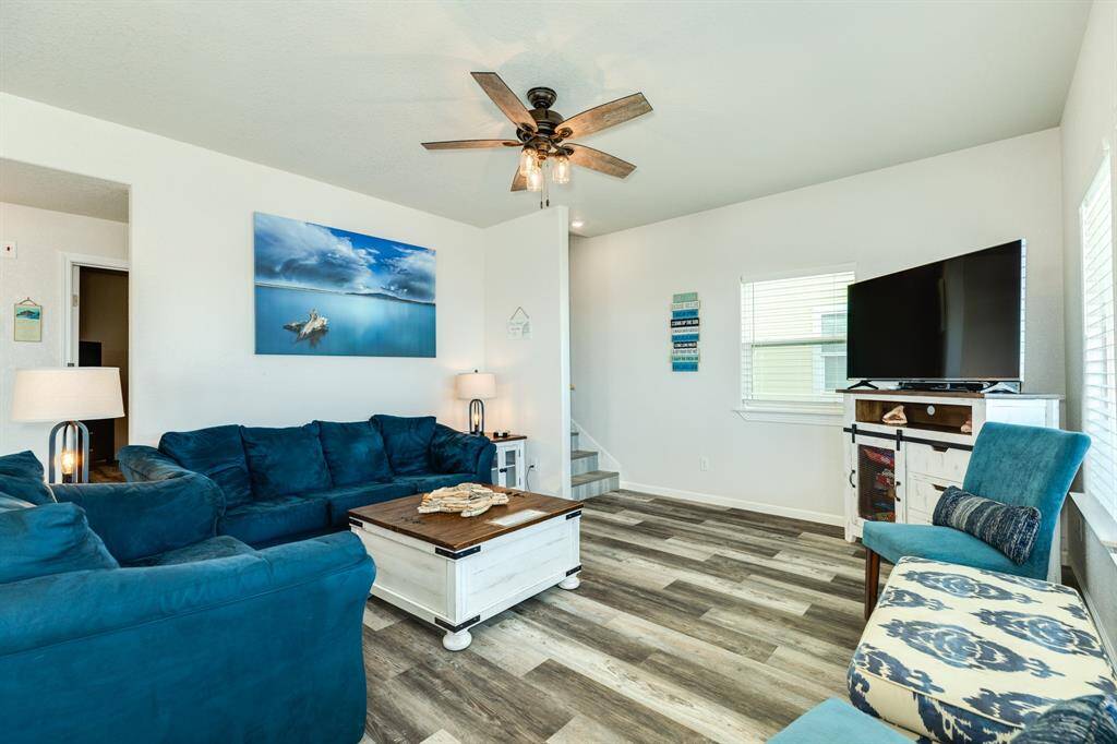 Bright and inviting living room with stylish flooring, a cozy blue sofa set, and a modern ceiling fan. Features a large window, wall art, and a TV, creating a comfortable space for relaxation and stairs leading to the second floor.