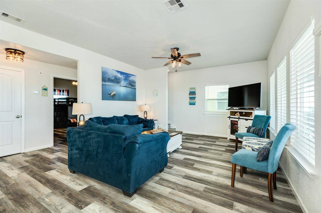 Bright and inviting living room featuring large windows, modern flooring, and a ceiling fan. The space is furnished with comfortable blue seating and a wall-mounted TV, offering a cozy atmosphere for relaxation.