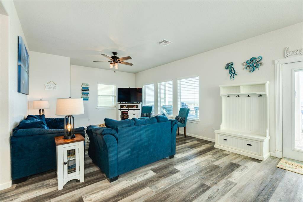 The living room features stylish flooring, a ceiling fan, and ample natural light from multiple windows. It is furnished with plush blue sofas, a TV setup, and a functional white coat rack with seating. The space has a coastal theme, perfect for relaxation.