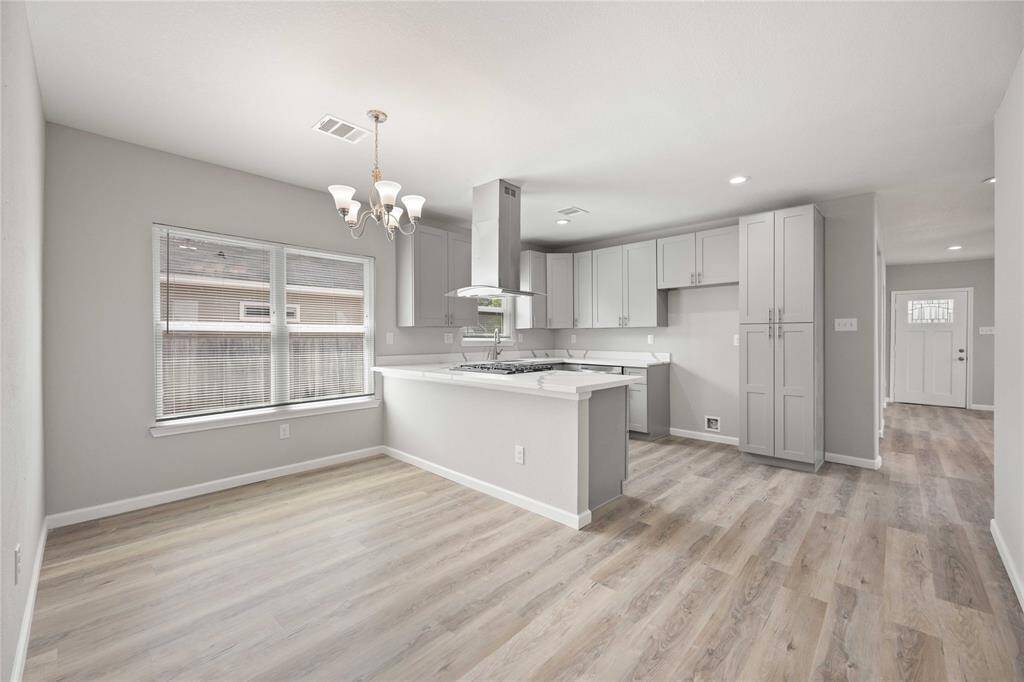 This photo showcases a modern, open kitchen and dining area with light-colored wood flooring and neutral-toned walls. The kitchen features sleek cabinetry, stainless steel appliances, and ample counter space. A large window provides natural light, enhancing the bright and airy atmosphere.