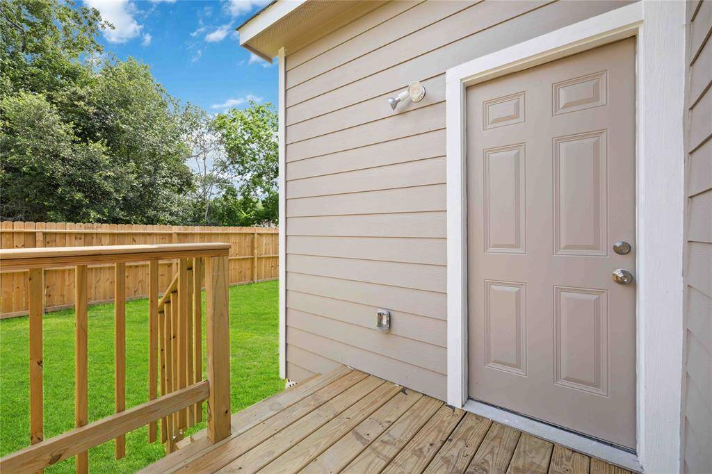 This photo features a cozy wooden deck with a light beige exterior door, leading to a fenced backyard with lush green grass and mature trees, perfect for outdoor enjoyment and privacy.