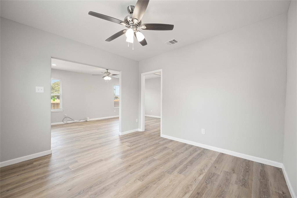 Bright and spacious room with modern light gray walls and wood-style flooring. Features ceiling fans and an open layout leading to another room, providing ample natural light.