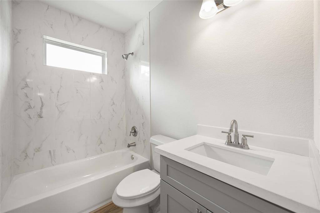 This bathroom features a modern design with a sleek vanity, white countertop, and a bathtub surrounded by elegant marble-style tile. A small window provides natural light, enhancing the bright, clean look.