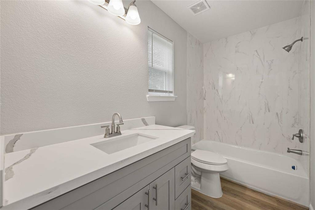 This bathroom features a modern design with a sleek, white vanity and marble countertop, complemented by a stylish bathtub with marble-patterned tile surround. It has a light, neutral color palette and wood-look flooring, creating a clean and inviting space.