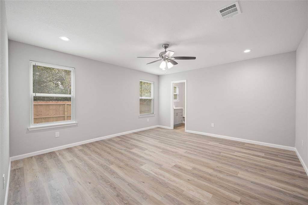 This spacious room features light wood flooring, neutral walls, and ample natural light from two windows. A ceiling fan provides comfort, and there's a doorway leading to an adjacent space, likely a bathroom or closet. Perfect for a bedroom or living area.