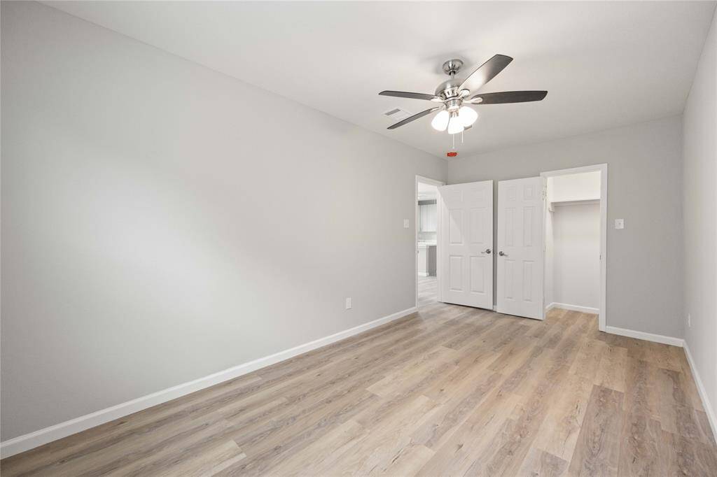 This is a bright, spacious bedroom featuring light gray walls, wood flooring, a ceiling fan, and a walk-in closet. It offers a clean, modern look with ample natural light.