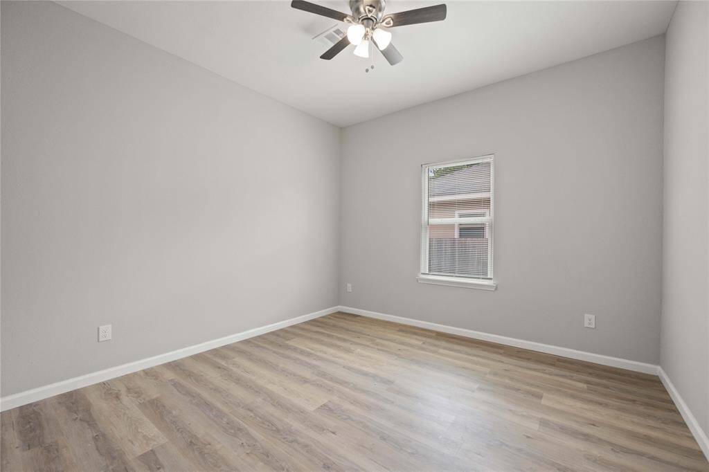 This room features light wood flooring, neutral gray walls, a ceiling fan with lights, and a single window with blinds, creating a bright and airy space.
