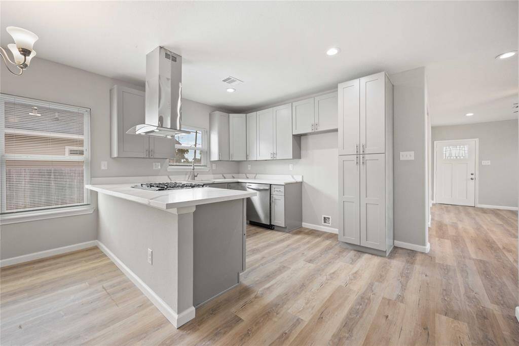 This photo showcases a bright, modern kitchen with light wood flooring, white cabinetry, stainless steel appliances, and a sleek gas cooktop on the island. The open layout and ample natural light create an inviting space.