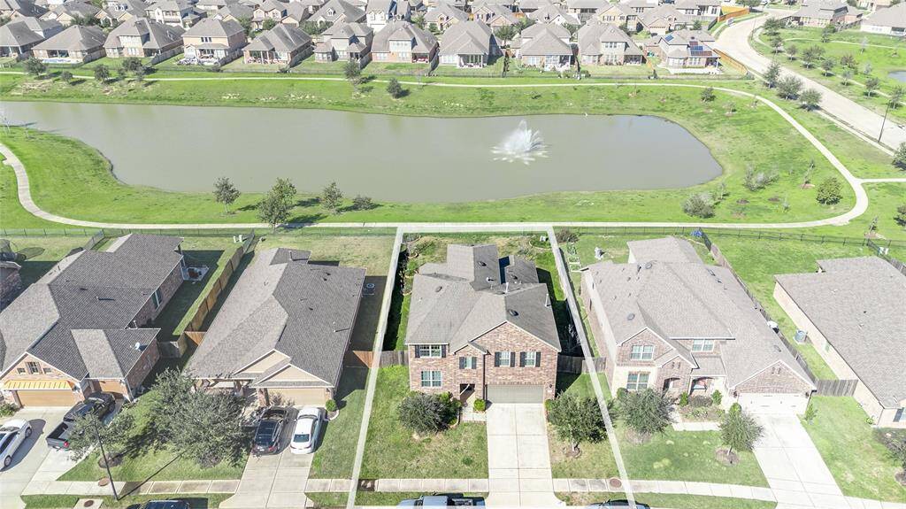 This aerial photo showcases a suburban neighborhood with a row of brick houses, each featuring a driveway and green lawns. Behind the houses is a scenic pond with a fountain, surrounded by a walking path and green space, offering a peaceful and attractive setting.