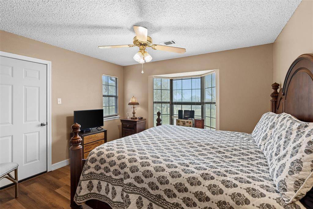 This cozy bedroom features a large bay window offering plenty of natural light and a ceiling fan. The neutral-toned walls and wood flooring create a warm atmosphere.