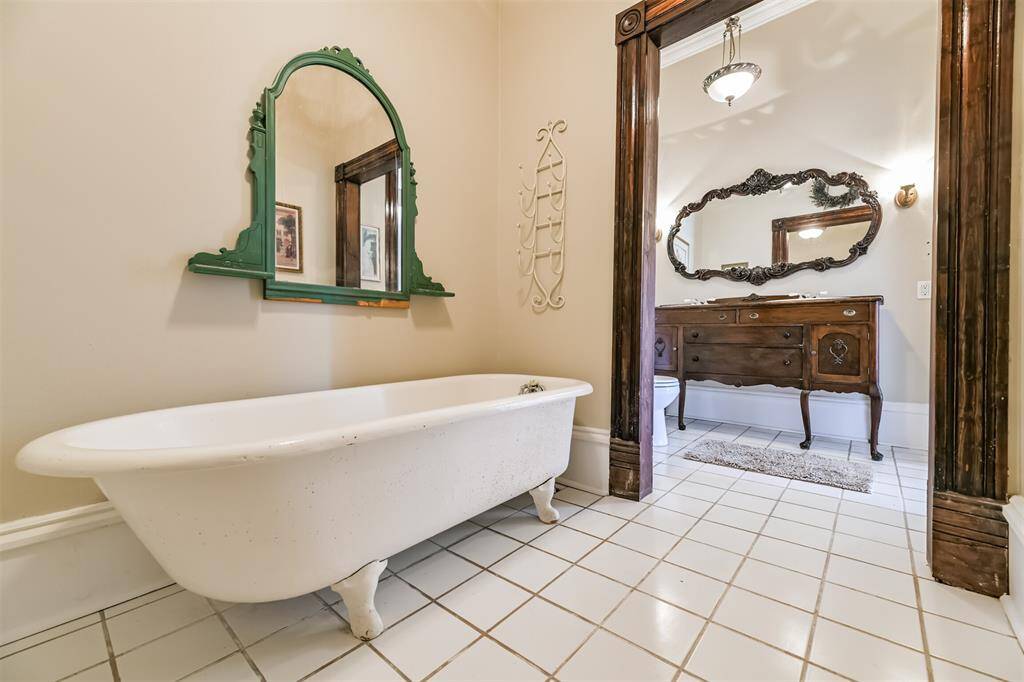 This charming bathroom services the secondary bedrooms, featuring a dresser-style vanity with dual basin sinks, a period-specific ornately framed mirror, tile floors, and a separate room with a cast iron clawfoot bathtub.
