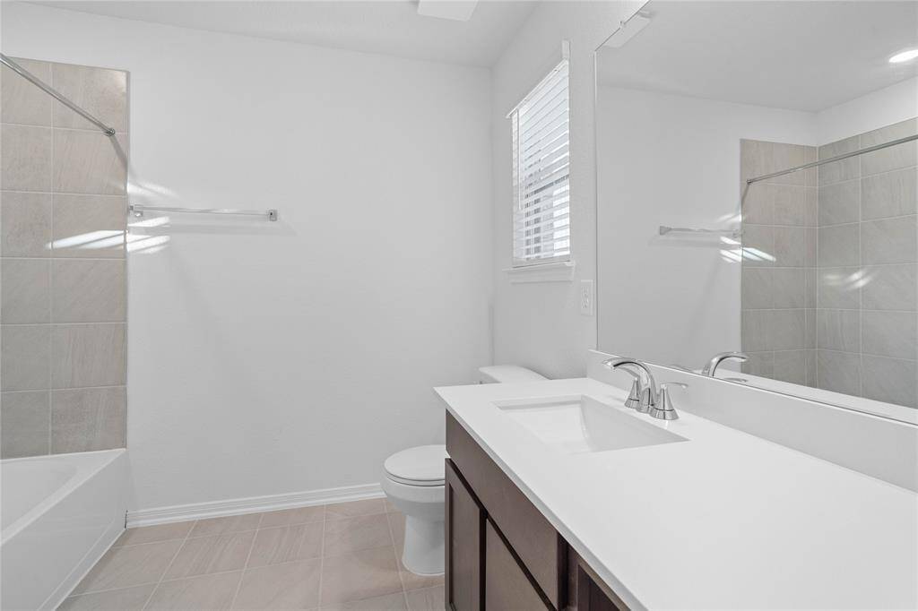Secondary bath features tile flooring, bath/shower combo with tile surround, window with blinds, stained wood cabinets, beautiful countertops, mirror, sleek fixtures and modern finishes.