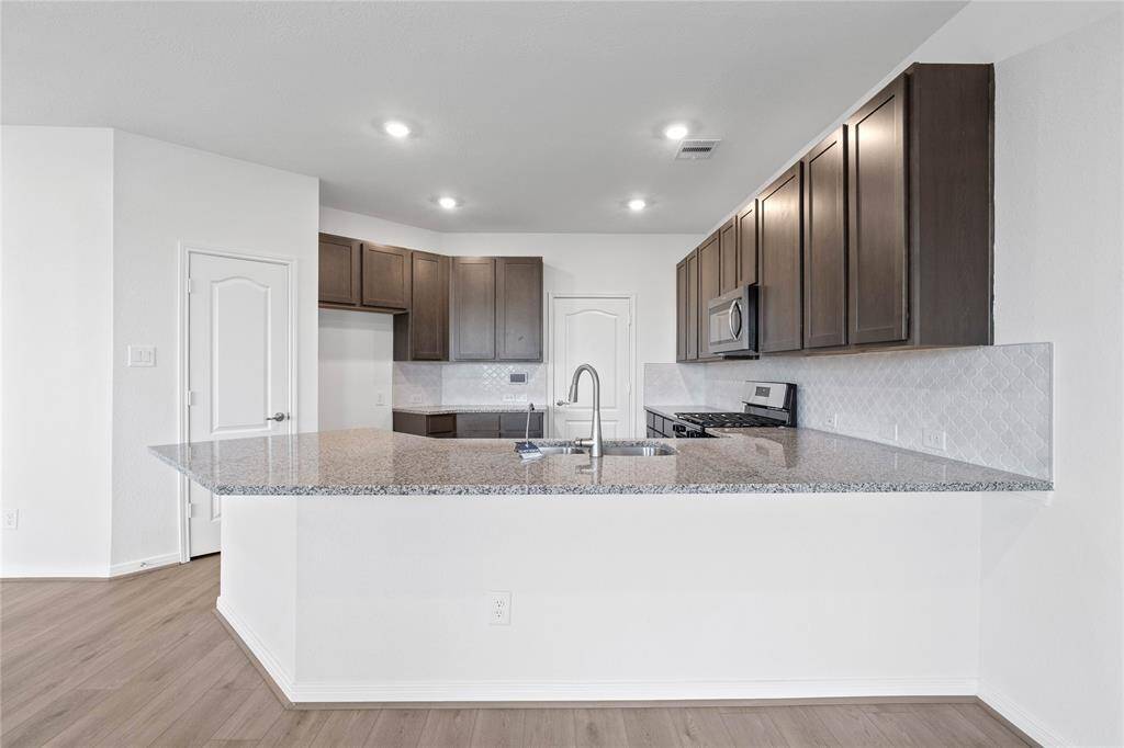 This spacious kitchen features high ceilings, recessed lighting, Woodmont Dakota cabinets in Smolder stain, granite countertops in Caramel Cream, SS appliances, modern tile backsplash, space for a breakfast bar, and a pantry all overlooking your huge family room.