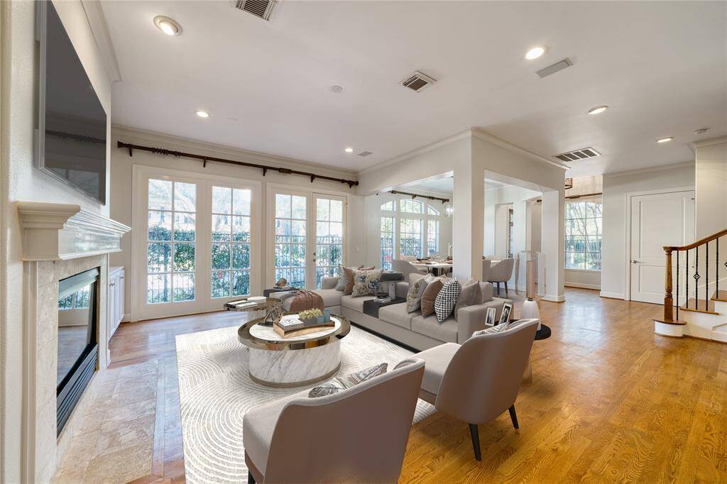An inviting view of the spacious living room and dining room in the distance, showcasing an open and airy entertaining space. This area is filled with natural light from a stunning wall of windows, complemented by recessed lighting throughout. A beautiful wrought iron staircase adds a touch of elegance, while the original wood floors enhance the charm and warmth of the home