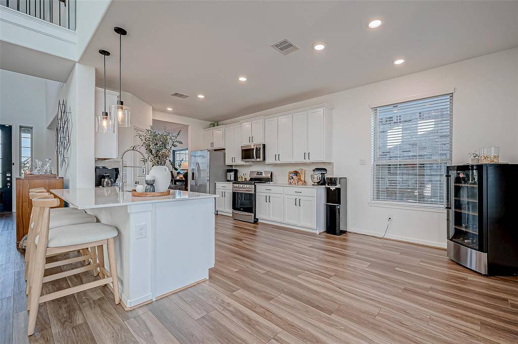 This modern kitchen features sleek white cabinetry, stainless steel appliances, and a spacious bar for seating. The open layout is complemented by laminate flooring and ample natural light from large windows, creating an inviting space for cooking and entertaining. Appliances: Refrigerator, Gas Range, Microwave. ( Water dispenser and wine refrigerator excluded)