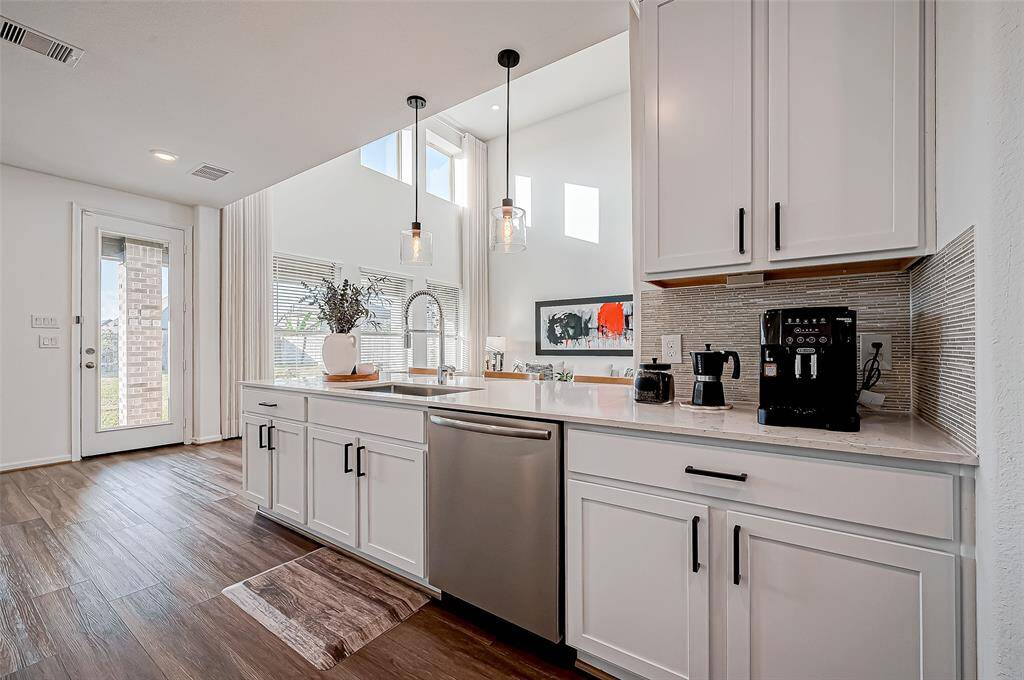 This modern kitchen features sleek white cabinetry, stainless steel appliances, and a stylish tile backsplash. It boasts ample natural light from large windows and high ceilings, with elegant pendant lighting above. The open layout is perfect for entertaining and offers easy access to the outdoor patio.