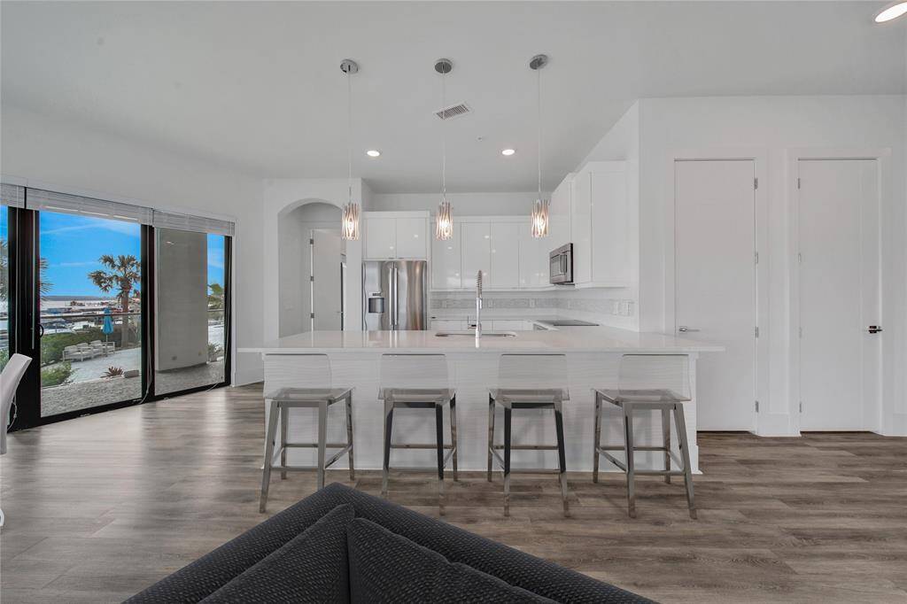 This open-plan kitchen and dining area with clean white finishes, stainless steel appliances, and a large island with seating. It features abundant natural light from sliding glass doors leading to an outdoor view.