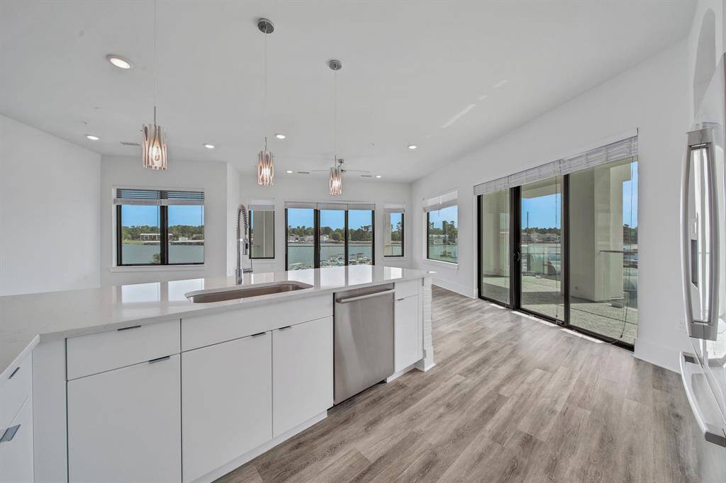 This bright kitchen with sleek white cabinetry and countertops, featuring elegant pendant lighting, and large windows with a view of the outdoors. It includes a spacious island and is equipped with contemporary appliances. The open-plan layout leads directly to an outdoor area, perfect for entertaining.