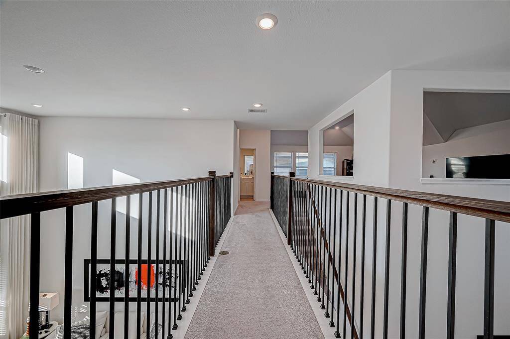 This photo shows a modern, carpeted upstairs hallway with black metal railing leading to the bedrooms and full bathroom. It overlooks a bright, open living space below, highlighted by large windows and recessed lighting. The second floors features 3 bedrooms, 2 full baths and game room.