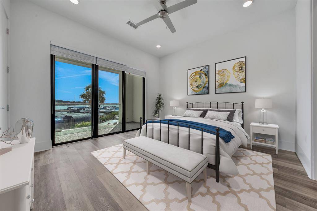 This Primary bedroom features large sliding glass doors that provide a stunning view of the outdoor landscape and water, allowing for plenty of natural light. The room is styled with contemporary furnishings and neutral tones, accented with blue for a calm and elegant aesthetic.