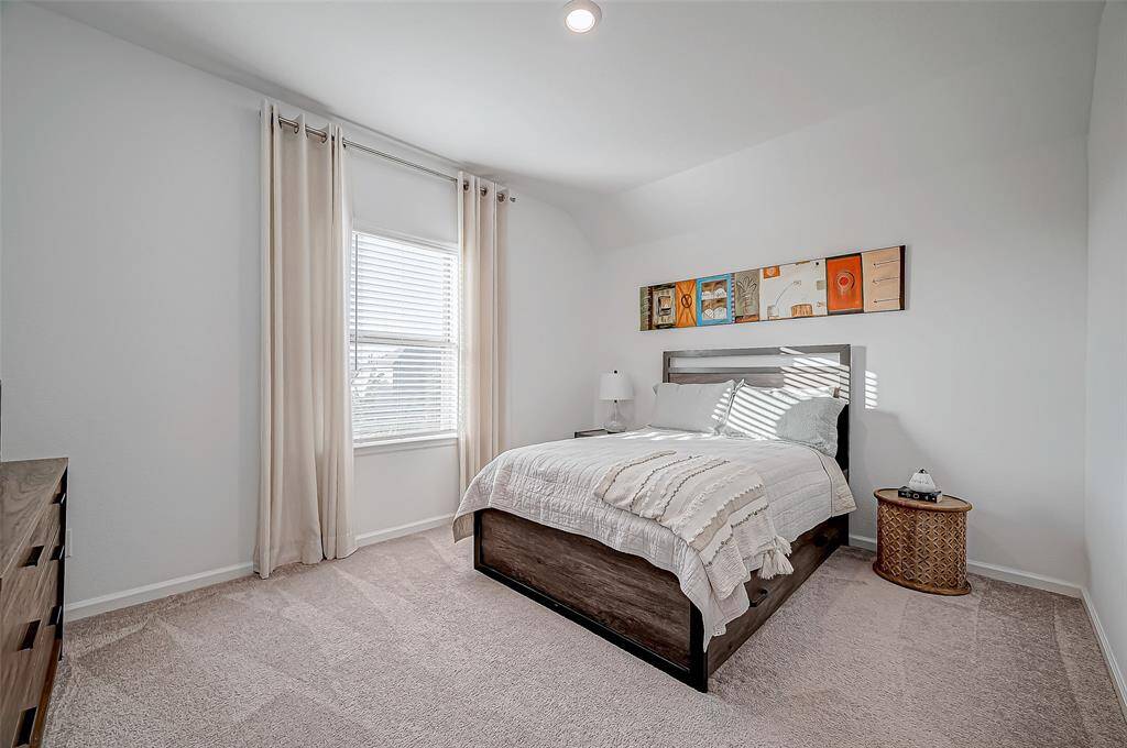 This cozy bedroom features soft carpeting, a large window with natural light, and neutral decor. The room features a queen bed, a nightstand with a lamp, and a dresser, creating a welcoming and functional space. (Furniture is not included in the sale)