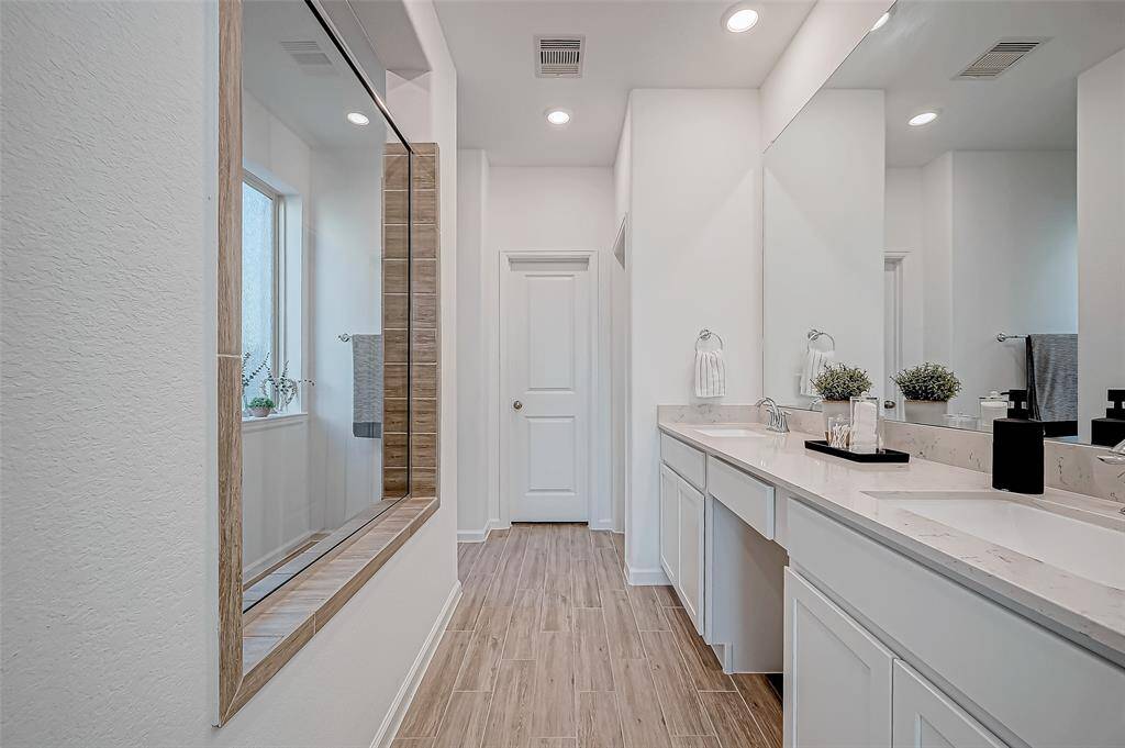 This photo showcases a bright, modern Primary bathroom with a spacious double vanity, sleek countertops, and ample storage. The wood-look tile flooring adds warmth, while the large mirror and recessed lighting enhance the room's airy feel. A window provides natural light, complementing the clean, neutral decor. (First Floor)