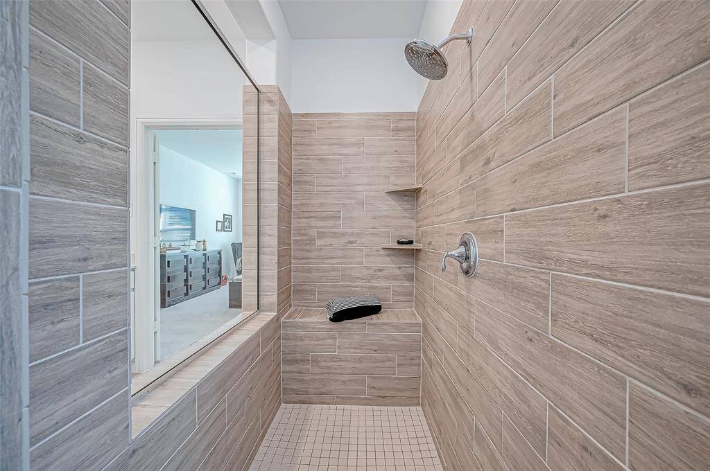 This photo showcases the primary bedroom modern and spacious shower with wood-look tile, sitting are, and a rainfall showerhead, providing a sleek and relaxing bathroom experience.