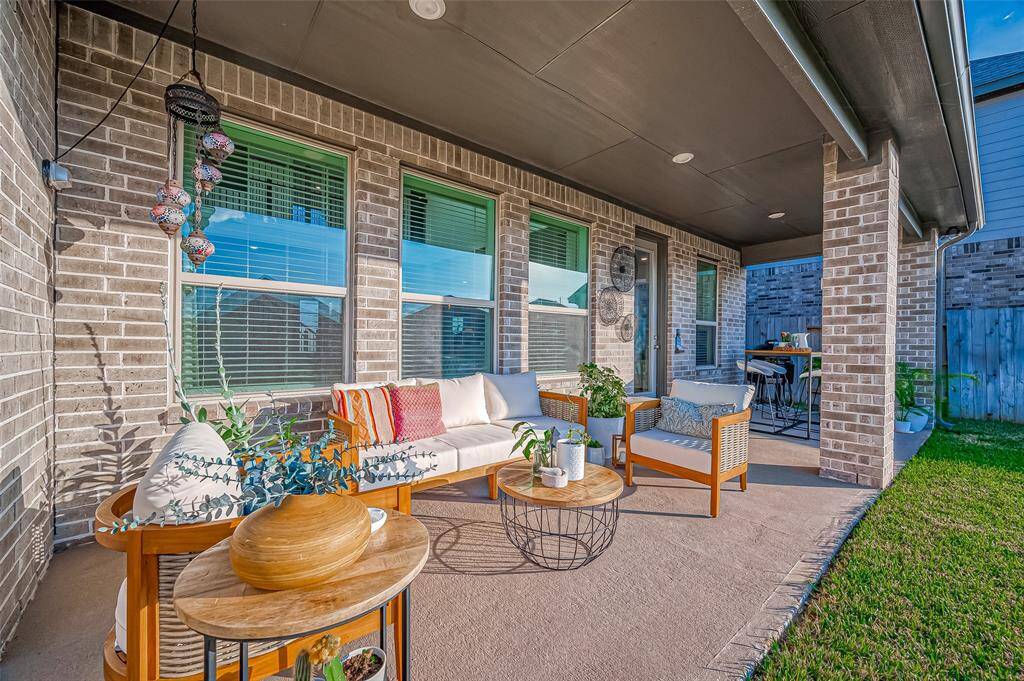 This photo showcases a cozy covered patio with stylish outdoor seating, a coffee table, and decorative plants. The space is perfect for relaxing and entertaining, featuring brick walls and a view of a small, well-maintained yard featuring sprinkler system front and back. ( All Patio furniture excluded)