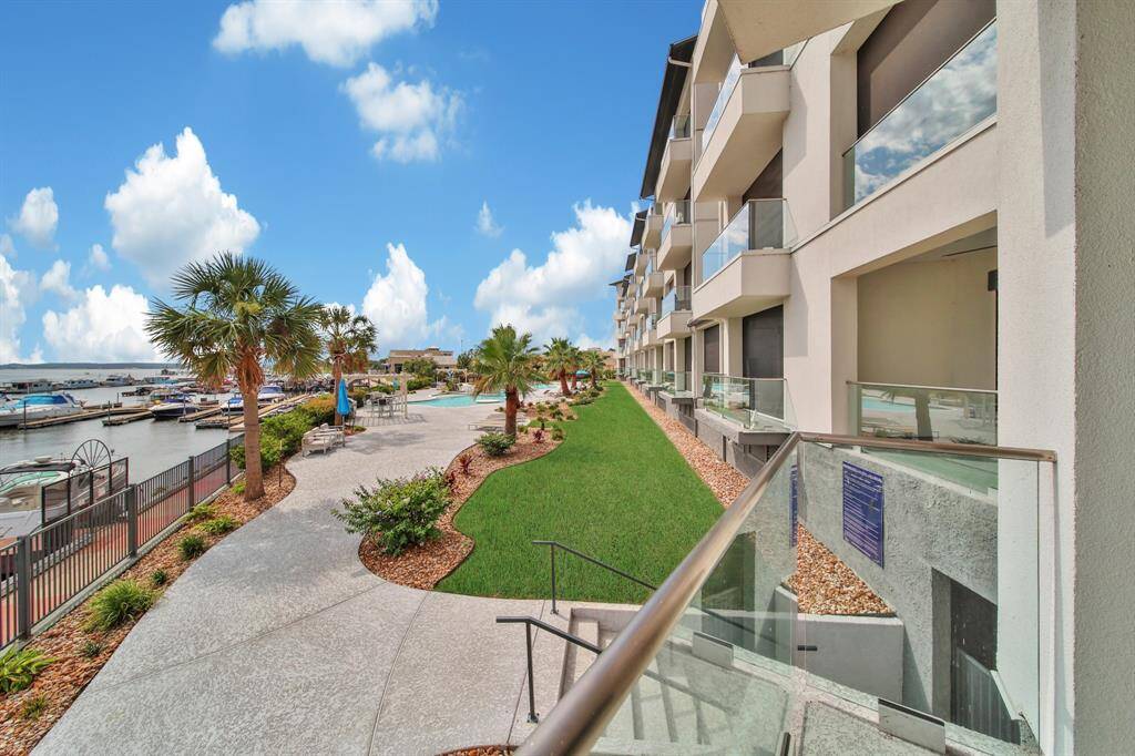 This is a waterfront condominium complex with a communal outdoor area, featuring a promenade with palm trees, seating, and views of a marina with boats. The building has multiple stories with private balconies, offering a modern living experience by the water.