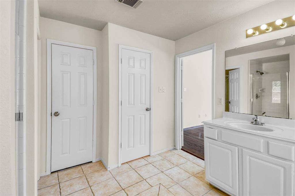 This is a bathroom featuring tiled flooring, white cabinetry with a sink, a large mirror with overhead lighting, and three white doors likely leading to a closet, toilet, and exit.