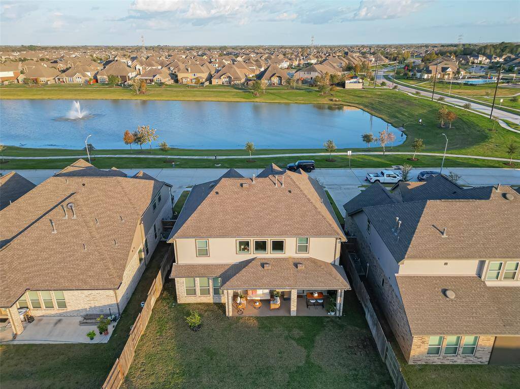 This aerial photo showcases a modern two-story home with a fenced backyard, situated in a suburban neighborhood. It offers a picturesque view of a nearby lake with a fountain,  providing a serene setting. The area is well-developed, with neighboring houses and accessible roads and parks. Short walking distance to the Community Pool.