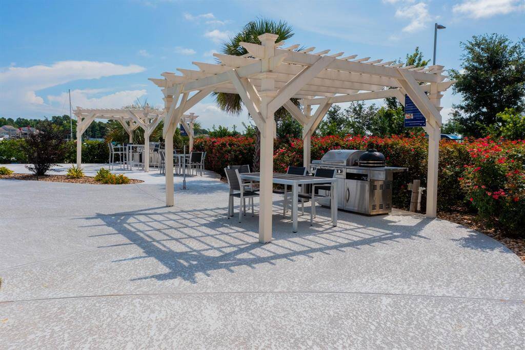 Amazing outdoor community space featuring a pergola with dining tables and a barbecue grill, surrounded by lush flowering bushes, ideal for entertaining and enjoying sunny days.