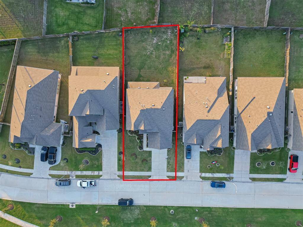 This aerial photo shows a row of suburban homes with well-maintained lawns. The highlighted property features a spacious backyard and a driveway leading to a two-car garage. The neighborhood appears quiet and neatly organized.