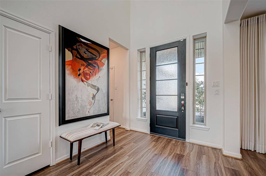 This photo showcases a modern entryway with a sleek black door flanked by tall windows. The space features laminate flooring, decorated with large abstract painting, and a stylish bench, offering a welcoming and artistic atmosphere.