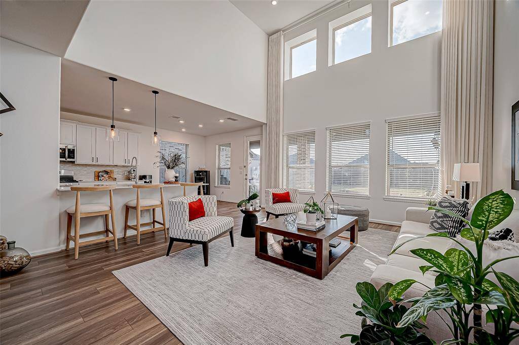 This photo showcases a spacious, open-concept living area with high ceilings and abundant natural light. The modern kitchen features a breakfast bar, and the living room is decorated with contemporary furniture and plants, creating a welcoming atmosphere. Custom Curtains & Rods in the living area will remain with the home.