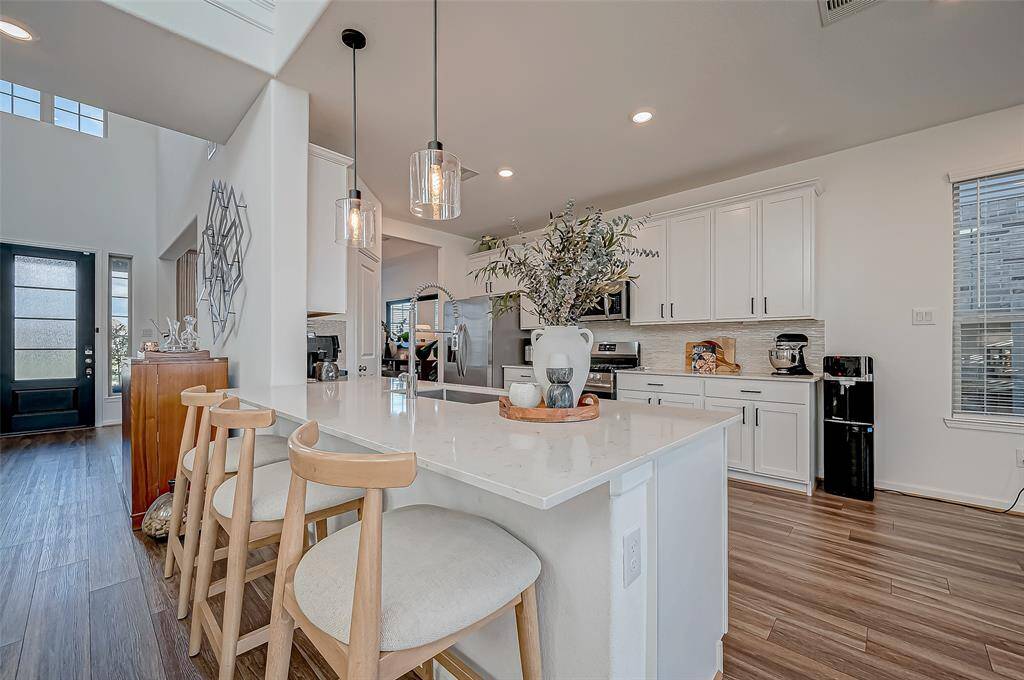 This modern kitchen features sleek white cabinetry, a spacious island with bar seating, and elegant pendant lighting. The open floor plan extends into a bright entryway with high ceilings and laminate floors, creating a welcoming and airy space.