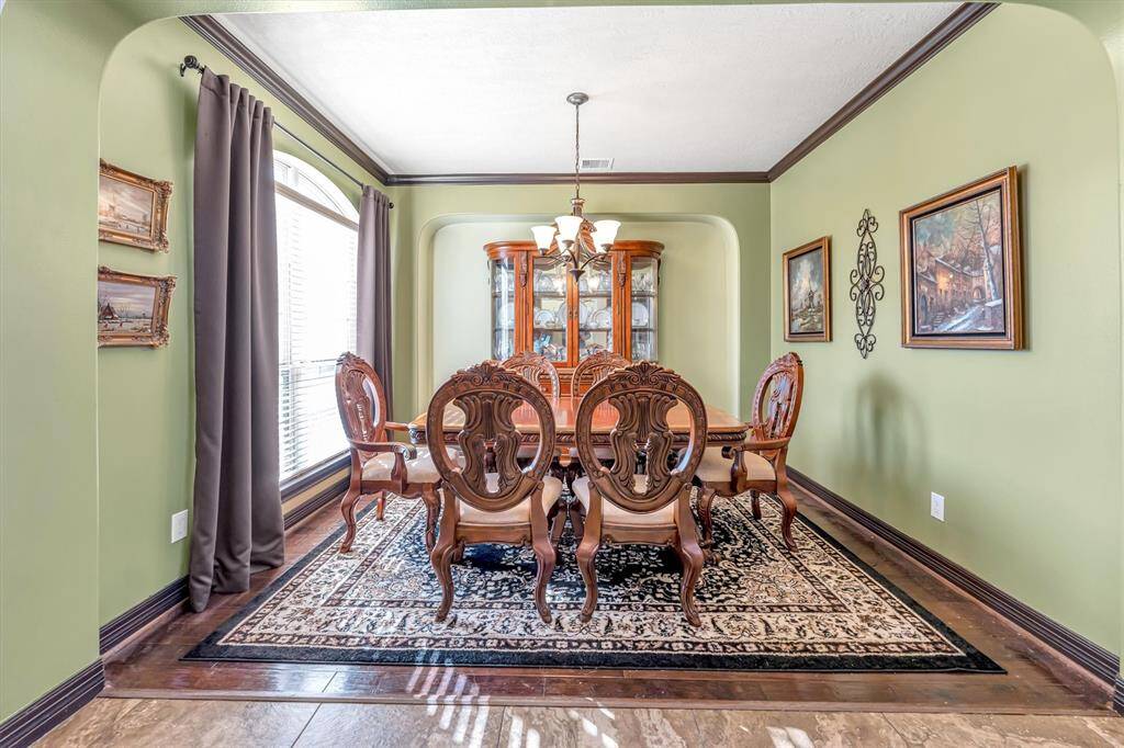 Discover a charming dining room featuring classic wooden furniture, a stunning chandelier, and elegant wall art. Natural light pours in through a large window with stylish curtains, enhancing the warm green walls and patterned rug on beautiful wood flooring.