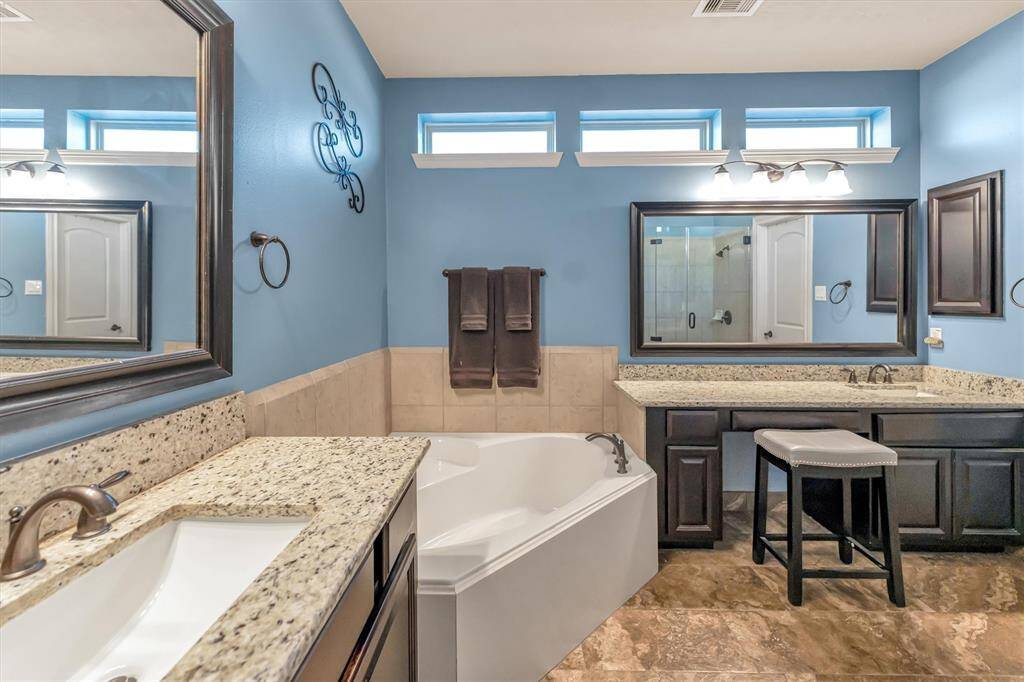 Spacious and modern primary ensuite bathroom featuring blue walls, granite countertops, and dark cabinetry. Enjoy a large soaking tub, a glass-enclosed shower, and dual sinks with stylish mirrors—perfect for relaxation.