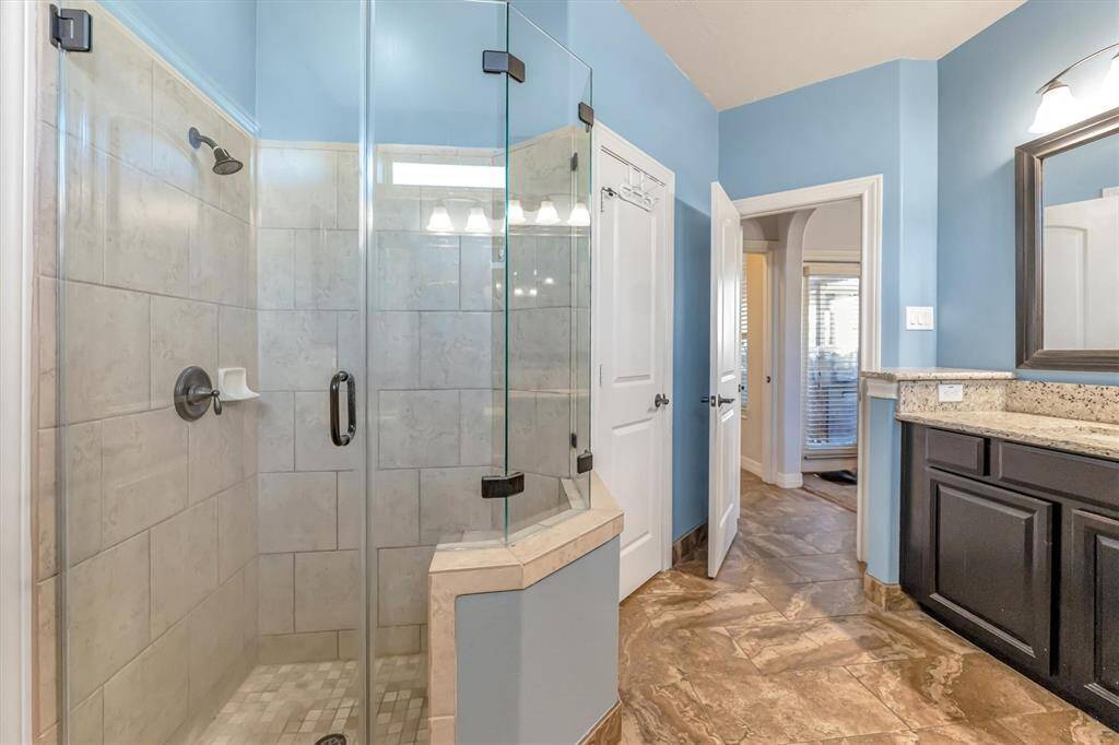 Ensuite bathroom boasts a spacious glass shower, elegant granite countertops, and rich dark wood cabinetry. The light blue walls and abundant natural light create a bright, airy atmosphere.