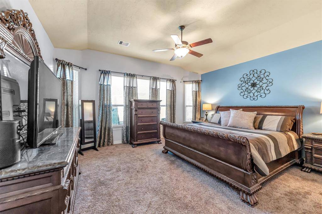 Inviting primary suite boasts a large bed, elegant wooden furniture, and abundant natural light. A ceiling fan and plush carpet add comfort, complemented by tasteful decor for a cozy ambiance.