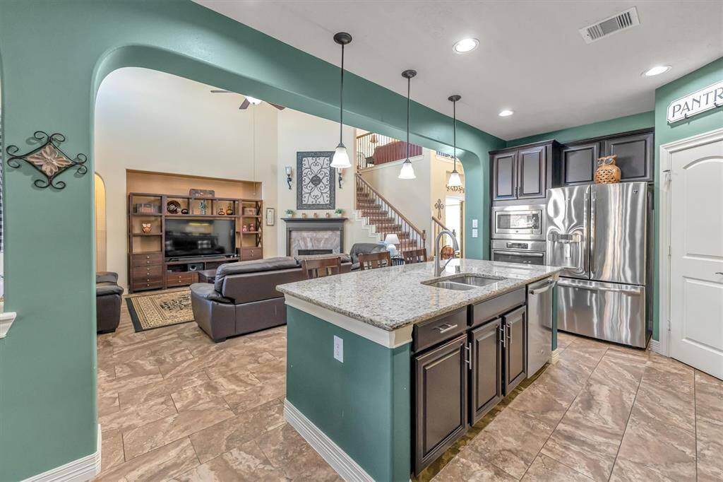 Discover a sleek, open-concept living area featuring a kitchen with granite countertops, stainless steel appliances, and rich cabinetry. The living room boasts high ceilings, cozy seating, and a built-in entertainment center, with a staircase enhancing the spacious ambiance.